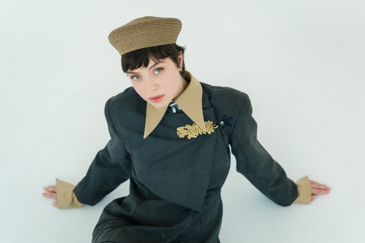 Editorial fashion portrait from above: a tailored charcoal jacket with gold cord and a woven beret, direct gaze