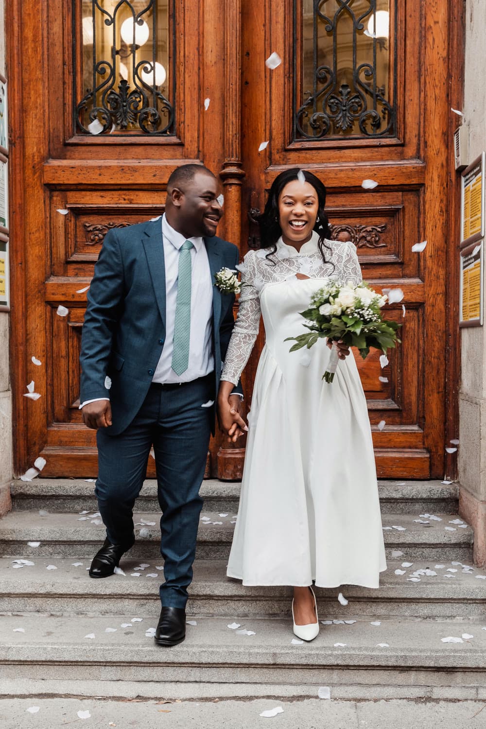A couple leaving a ceremony hand in hand under falling confetti, bouquet in hand